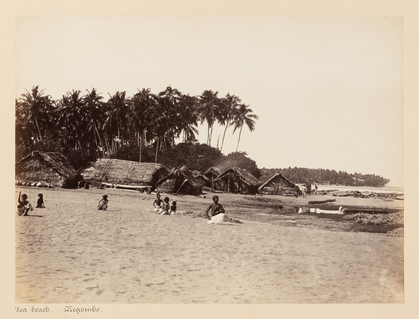 View of a beach showing people with palm trees and huts in the background, Negombo, Ceylon (now Sri Lanka)