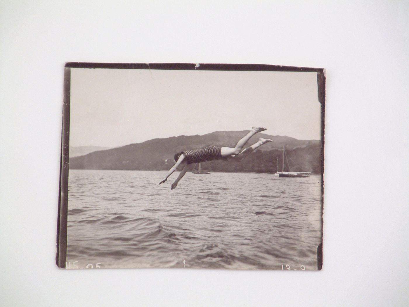 View of a man in one-piece swimsuit diving into water, with hills and sailboats in the distance, near Zambezi River