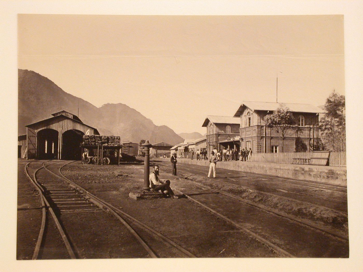 View of the Orizaba railway station showing men standing on the platform with a train shed on the left and mountains in the background, Mexico
