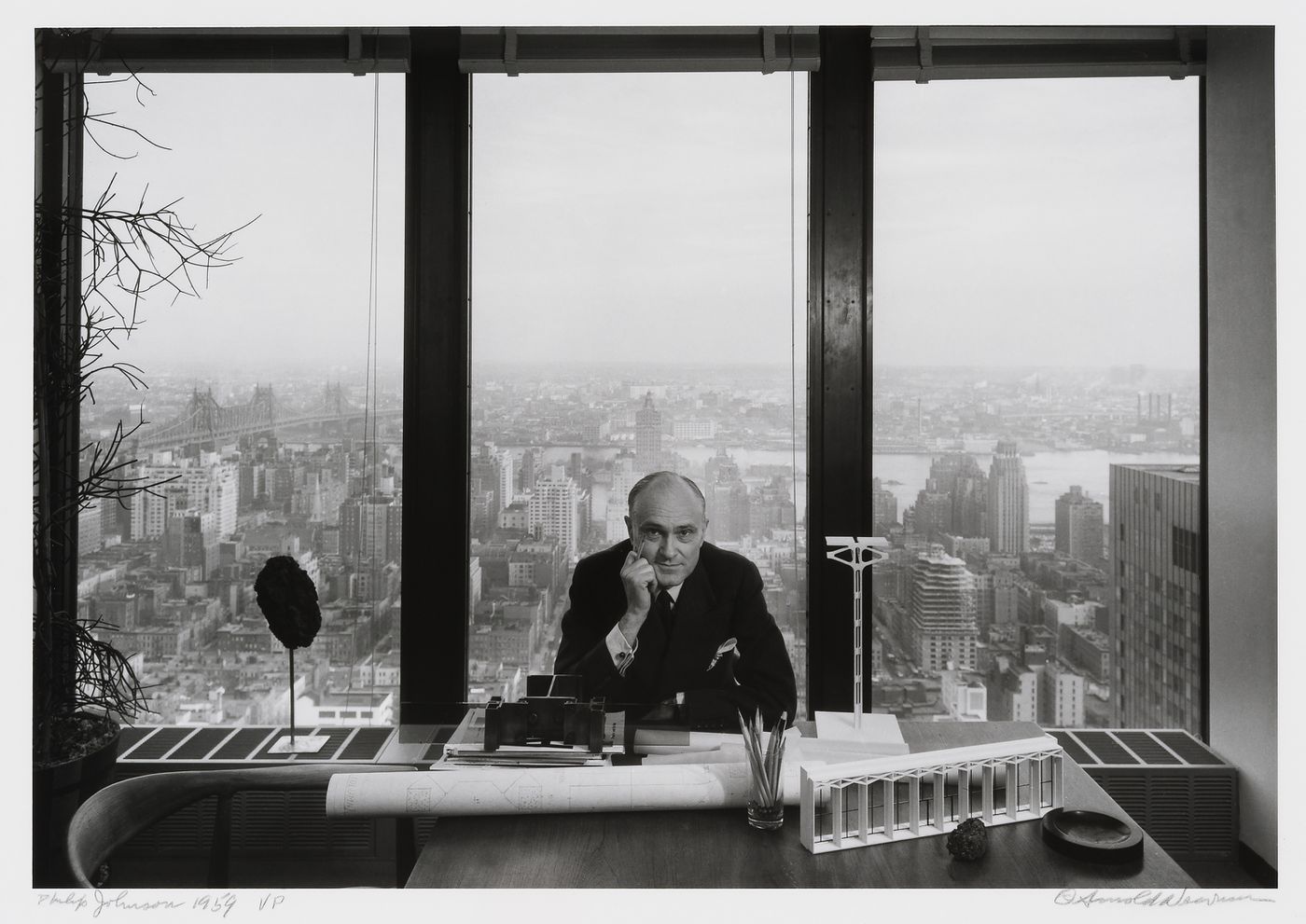 Portrait of Philip Johnson at his desk in the Seagram Building with a view of buildings and the East River in the background, New York City