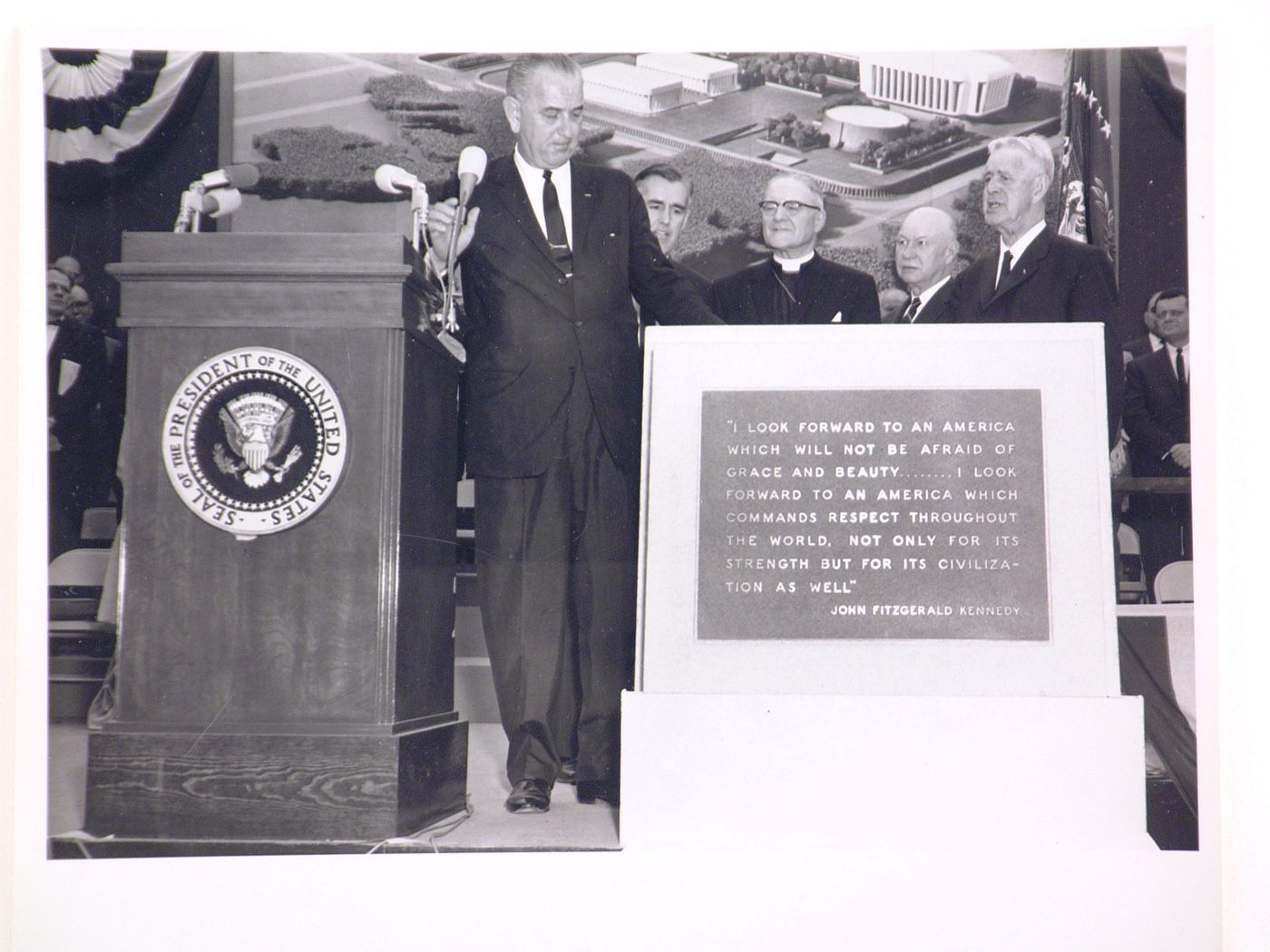 View of the dedication ceremony of the John F. Kennedy Educational, Civic and Cultural Center showing the memorial plaque with a quote by John F. Kennedy with President Lyndon B. Johnson and other men of state on the platform, Mineola, New York