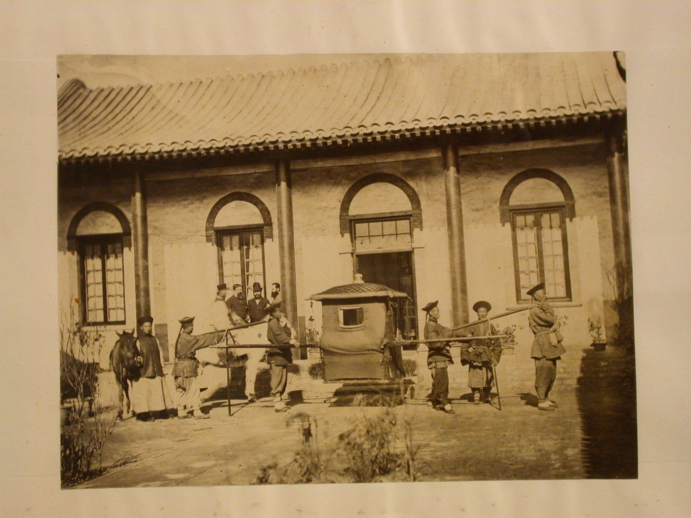 View of a litter with porters in front of a Chinese building with Westerners waiting, Peking (now Beijing) ?, China
