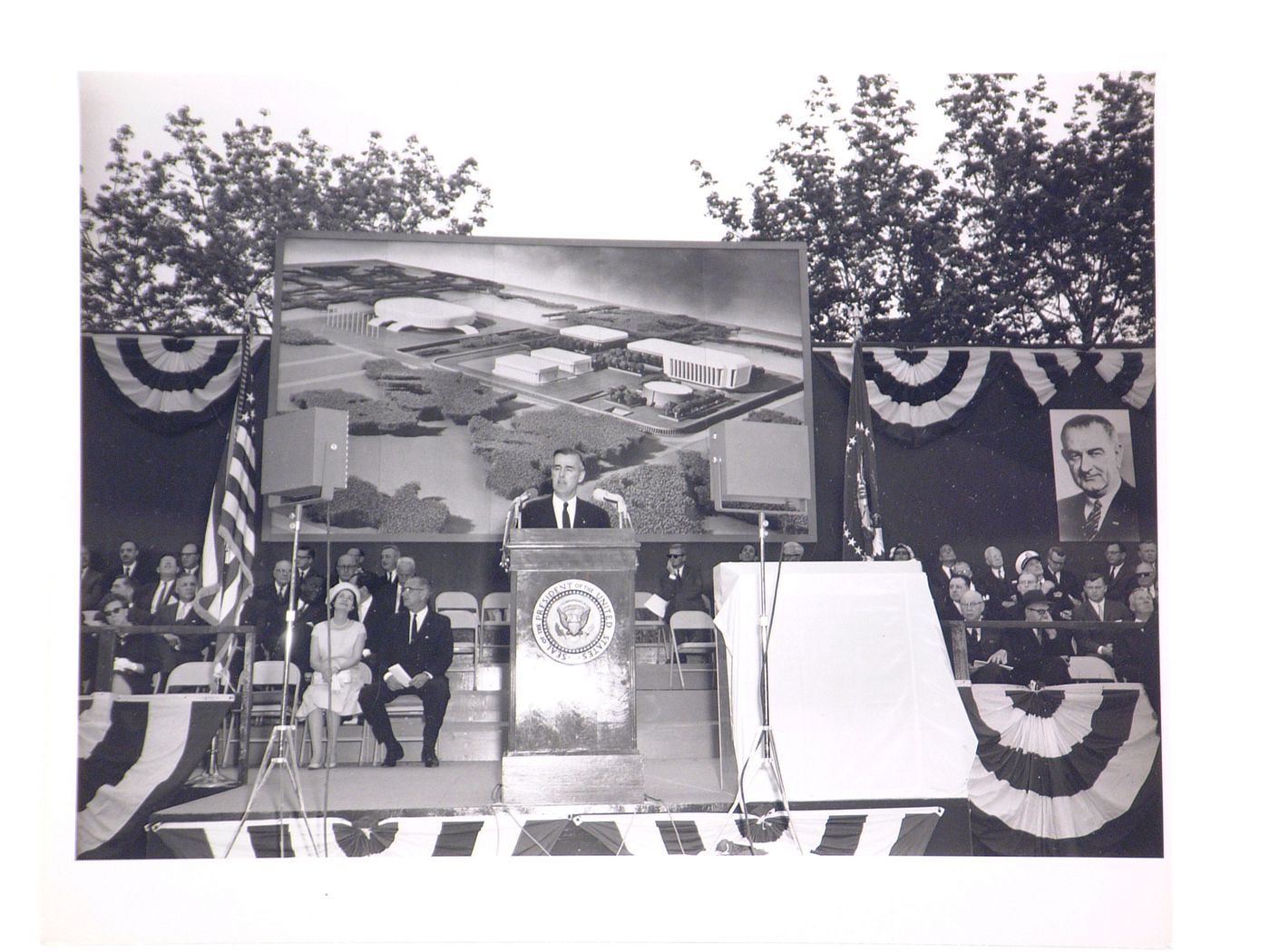 View of the dedication ceremony of the John F. Kennedy Educational, Civic and Cultural Center showing a partial view of the platform with people, posters of President Lyndon B. Johnson and a photograph of the model of the building, Mineola, New York
