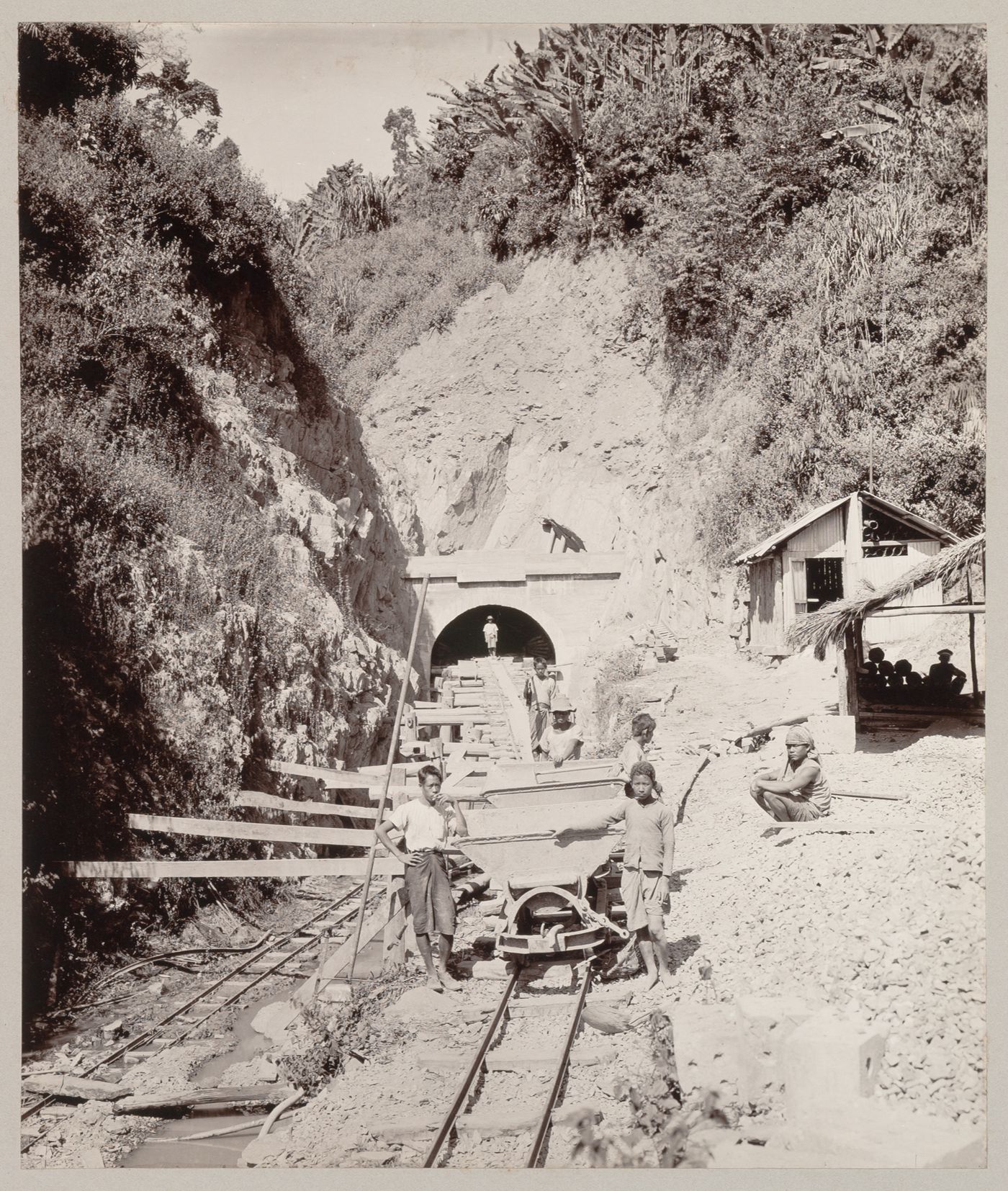 View of two smoking workers and other workers, hopper cars, tracks and the entrance to a railroad tunnel under construction, probably Khao Phlung hill, Phrae Province, Siam (now Thailand)
