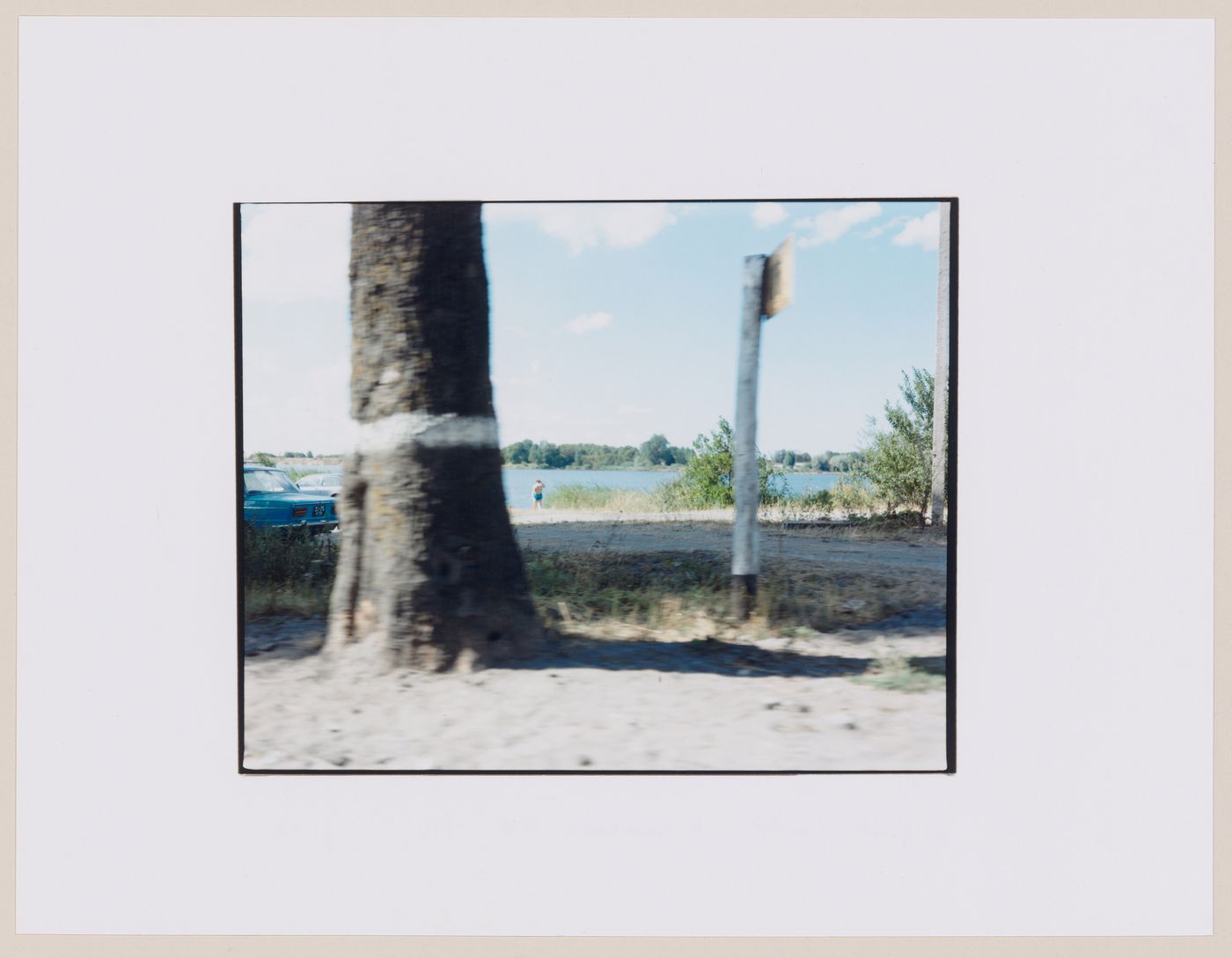 View of the entrance to a beach showing a tree trunk, a guidepost, automobiles, a woman and a body of water, Mamonovo, Kaliningradskaia oblast', Russia (from the series "In between cities")