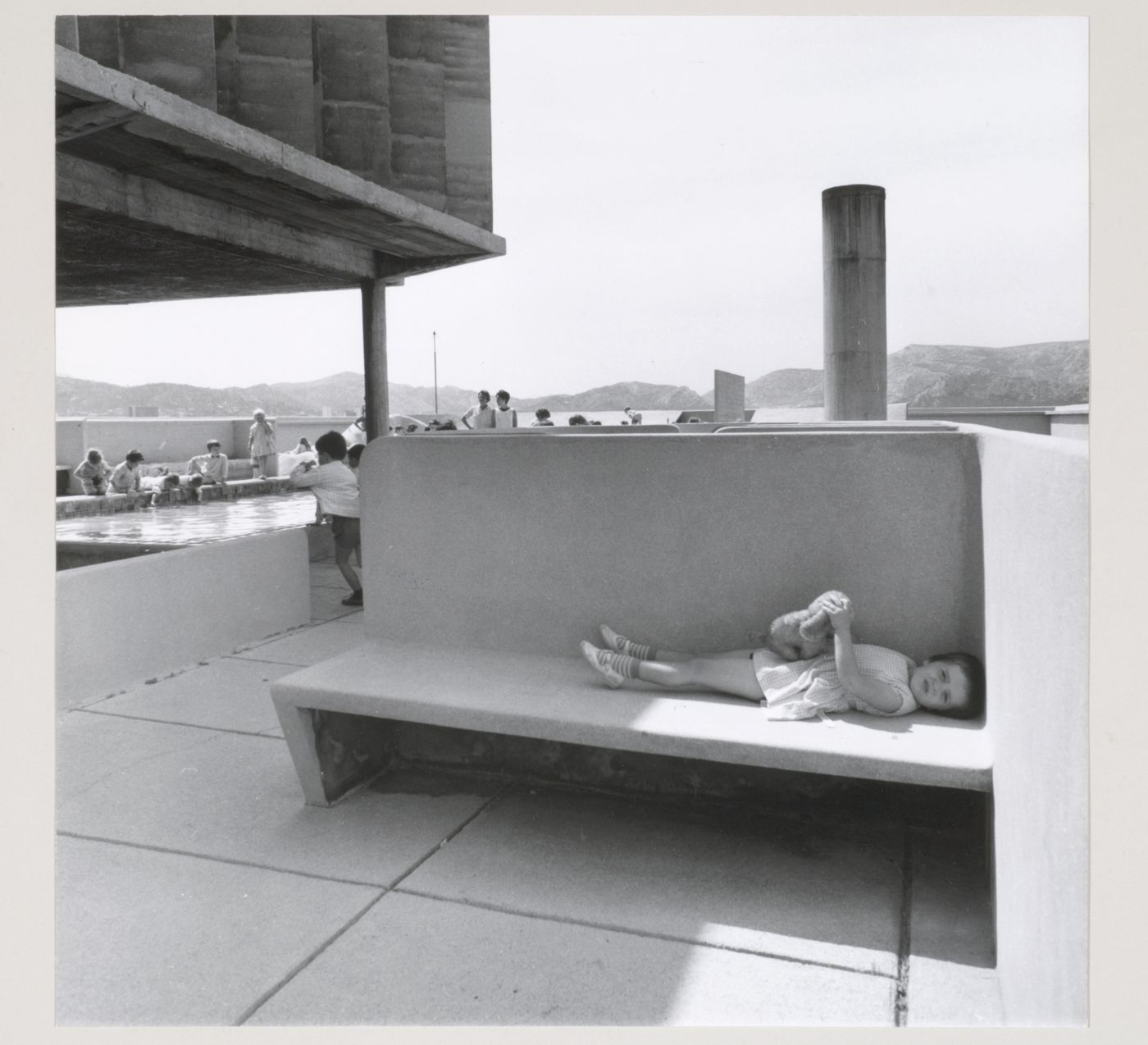 View of the roof of Unité d'habitation showing a girl lying on a concrete bench with children playing by the pool in the background, Marseille, France