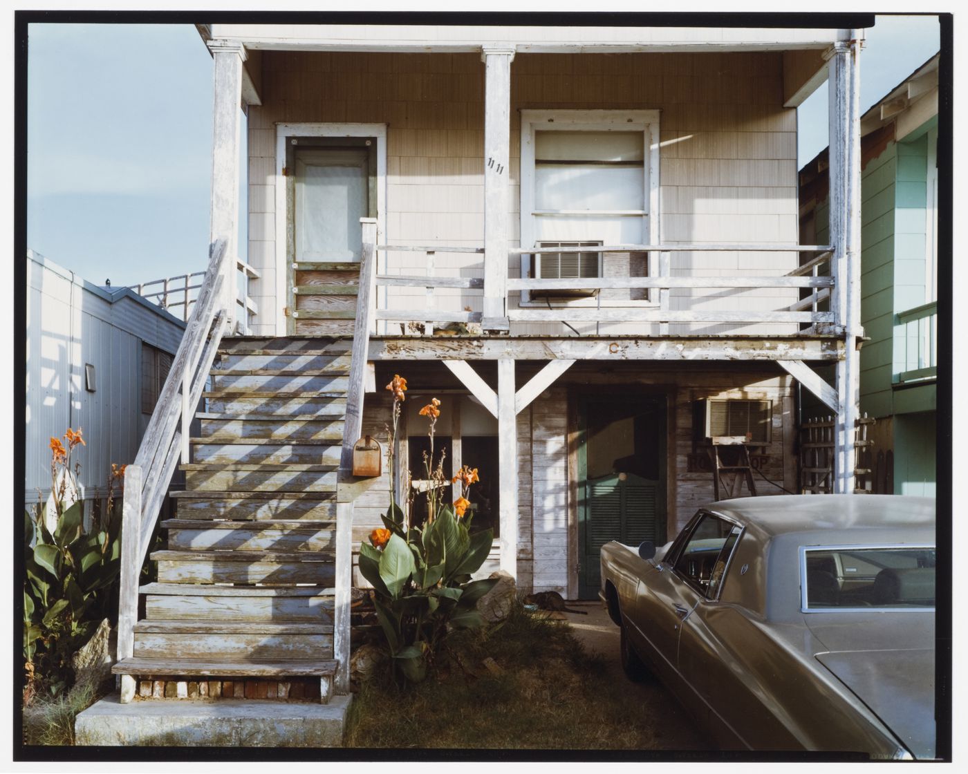 View of house with stairs leading up to a porch, flowering plants and a car, M 1/2 Ave., Galveston, Texas