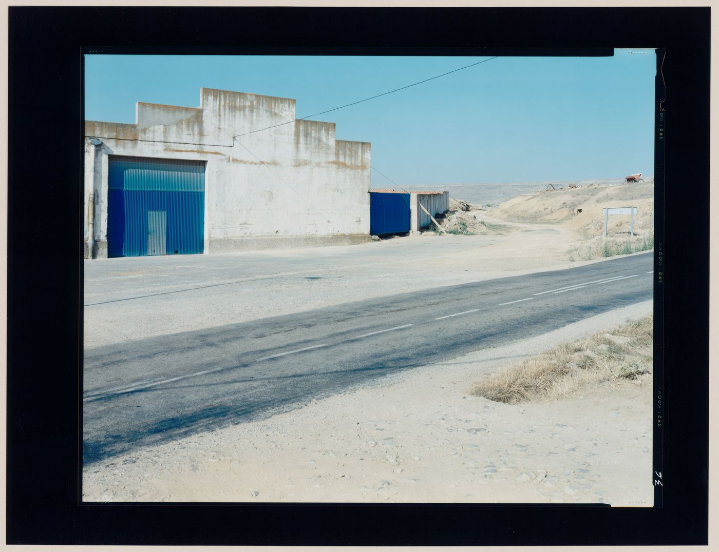 View of an agricultural building showing a concrete wall, a metal door, a road and hills, Castrojeriz, Burgos Province, Spain (from the series "In between cities")