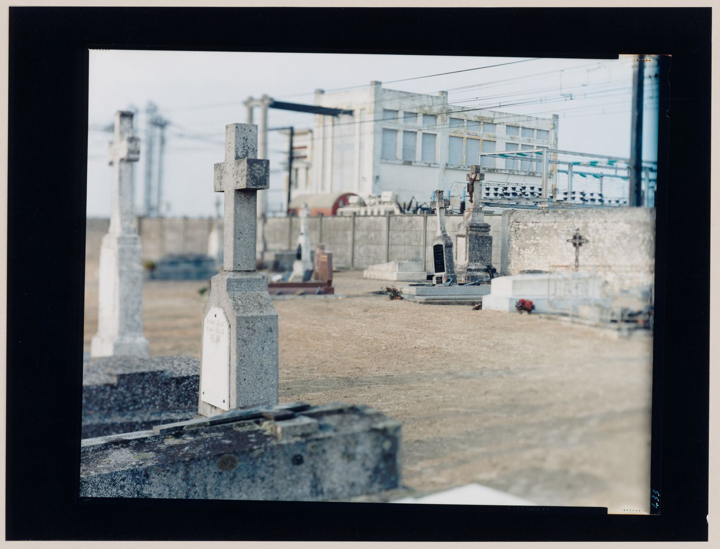 View of tombstones and a cemetery wall showing power lines and a building in the background, Petit Niort, near Saintes, France (from the series "In between cities")