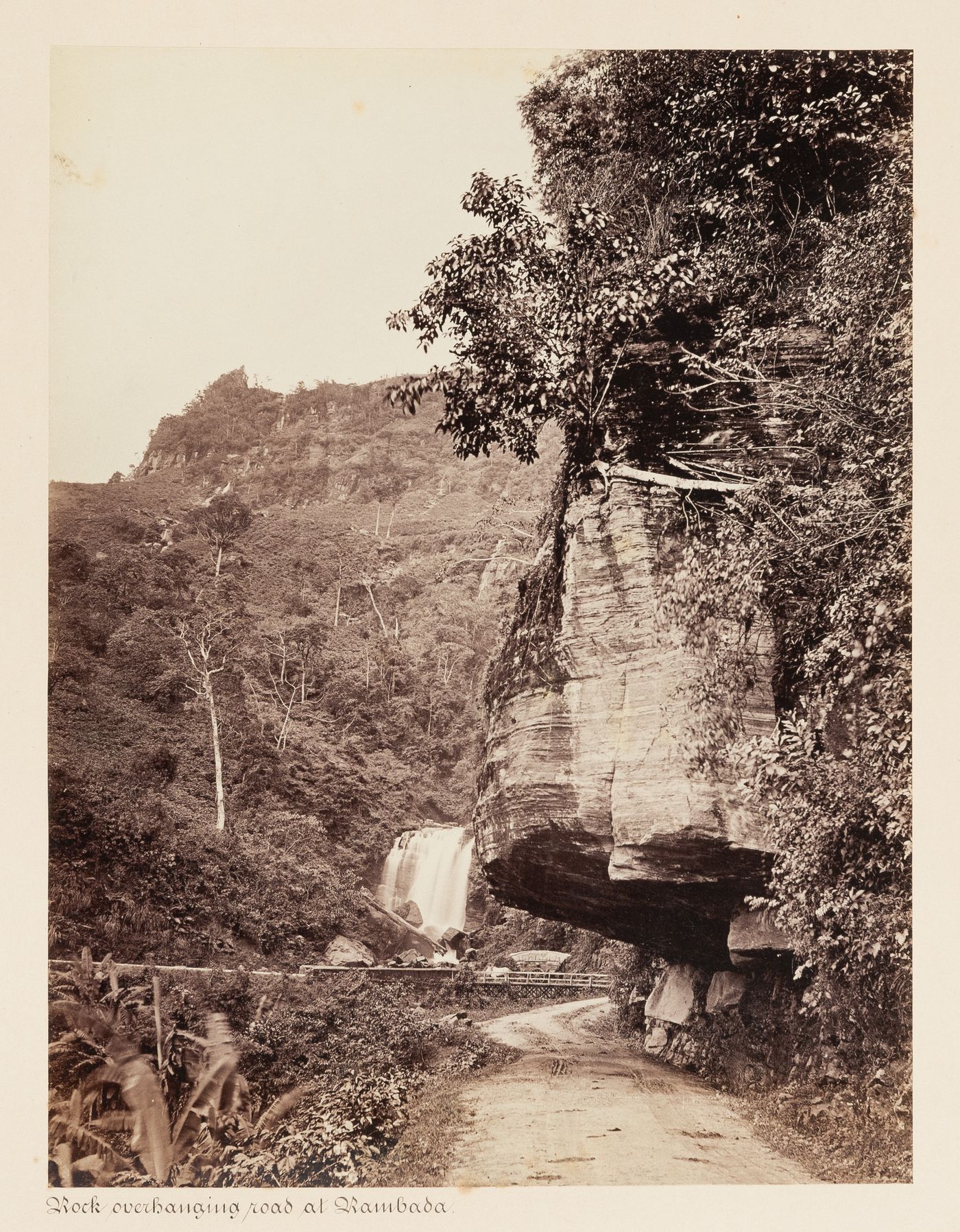 View of rock overhanging a road with the Wavedon Falls in the background, Ramboda, Ceylon (now Sri Lanka)