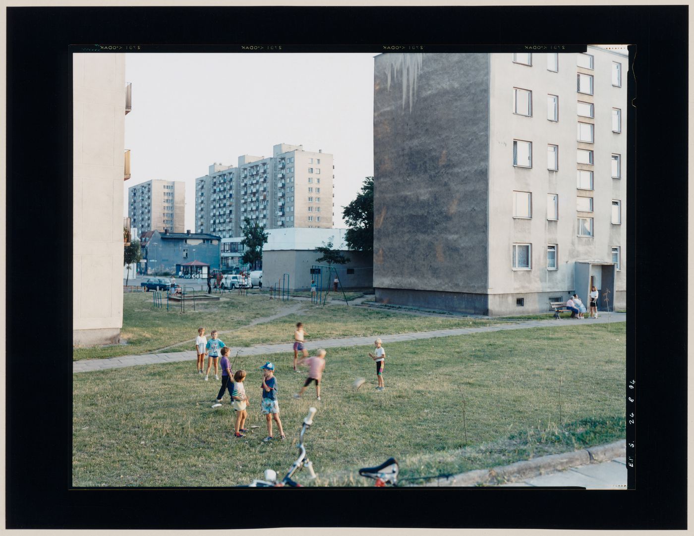 View of apartment houses and children playing on a lawn showing a playground in the background, Malbork, Poland (from the series "In between cities")