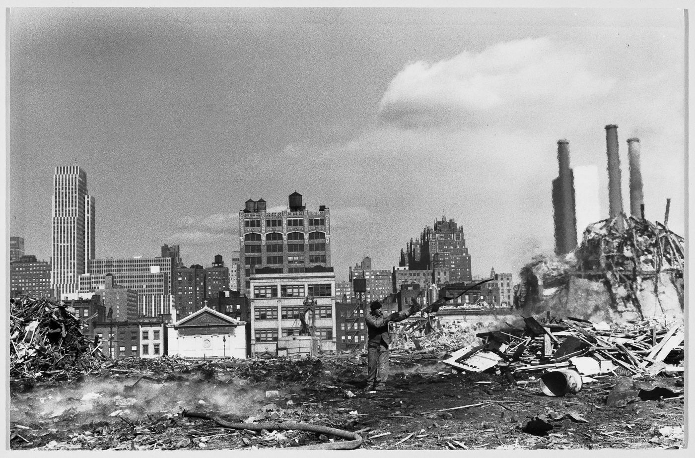 View of a demolition site [?] showing a man and fire hose with skyscrapers and smokestacks in the background, New York City, New York