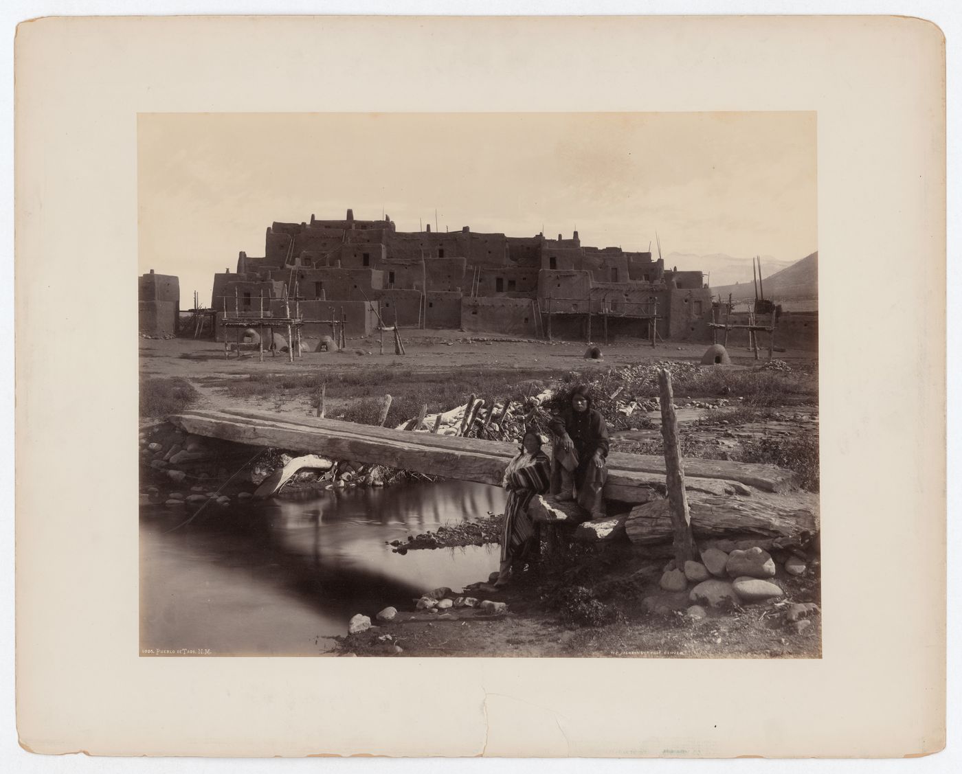 View of Taos Pueblo with two possibly Taos people in front of wooden bridge in the foreground, New Mexico, United States