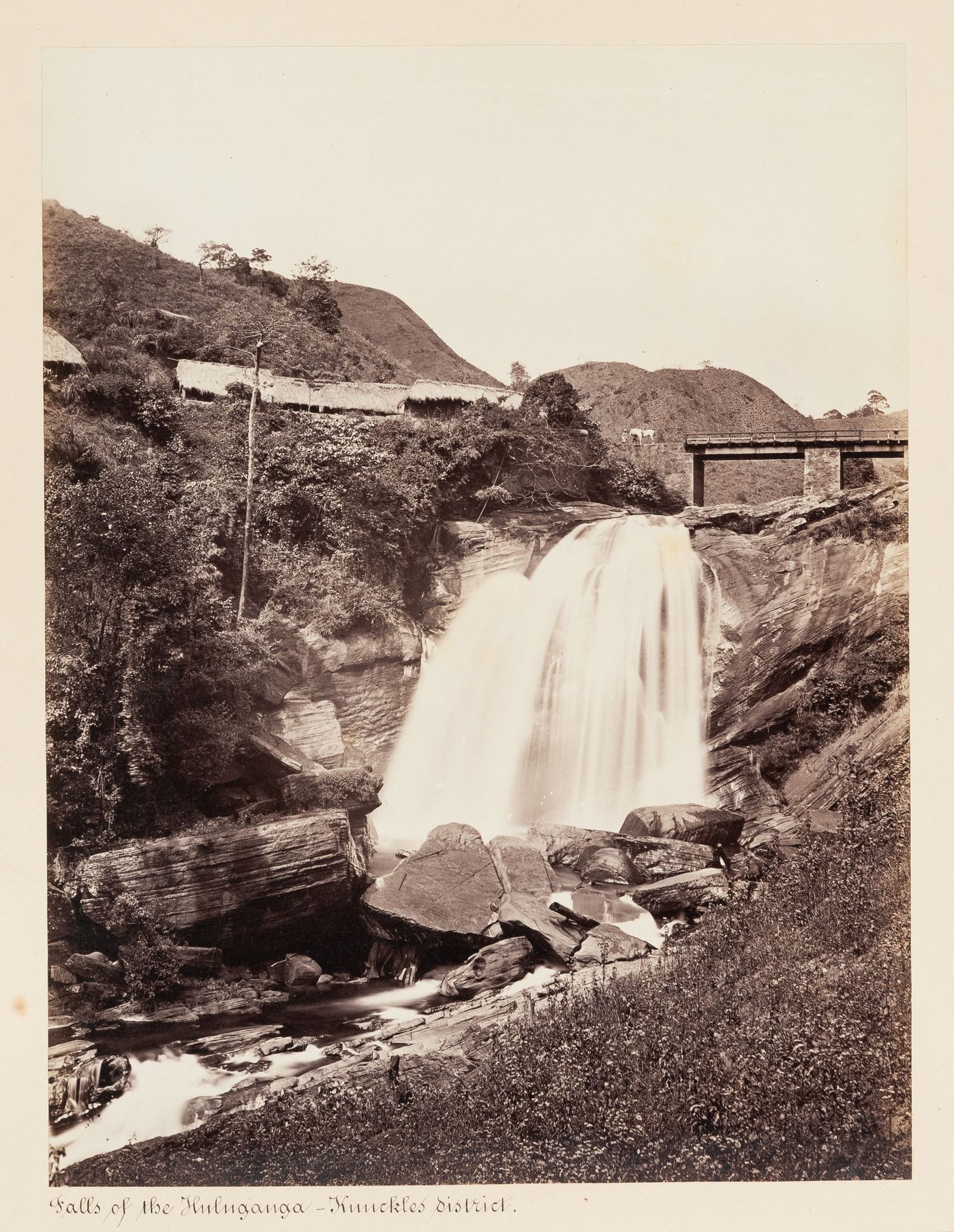 View of the Falls of the Hulu Ganga showing buildings and a bridge, Knuckles District [?], Ceylon (now Sri Lanka)