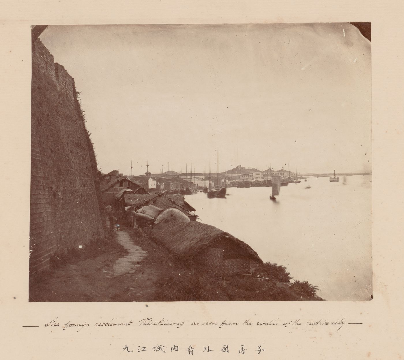 View of thatched buildings on river bank outside city wall with port and the British Concession in the background, Kiukiang (now Jiujiang), China