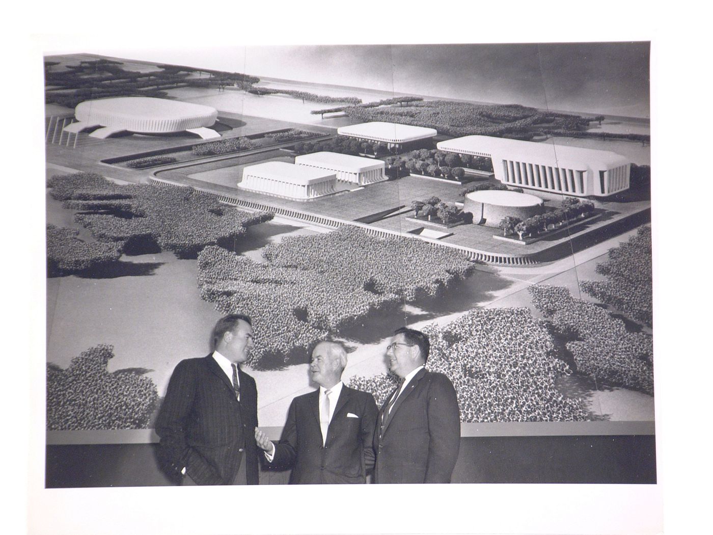 View of the dedication ceremony of the John F. Kennedy Educational, Civic and Cultural Center showing a photograph of the model of the building and three men on the platform, Mineola, New York