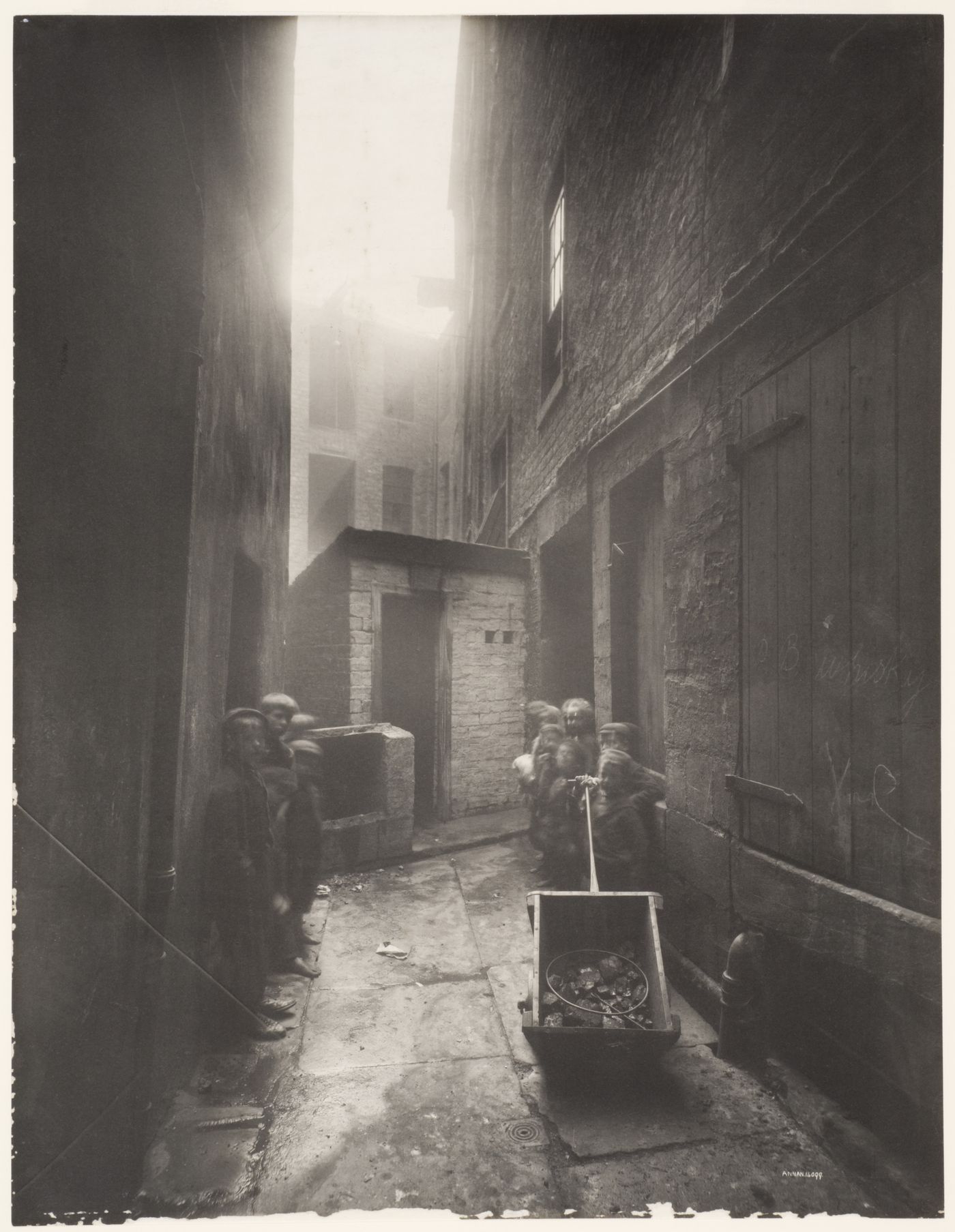 View of a narrow alley leading to a brick shed before which are many children, some pulling a wheeled cart containing coal, Glasgow, Scotland