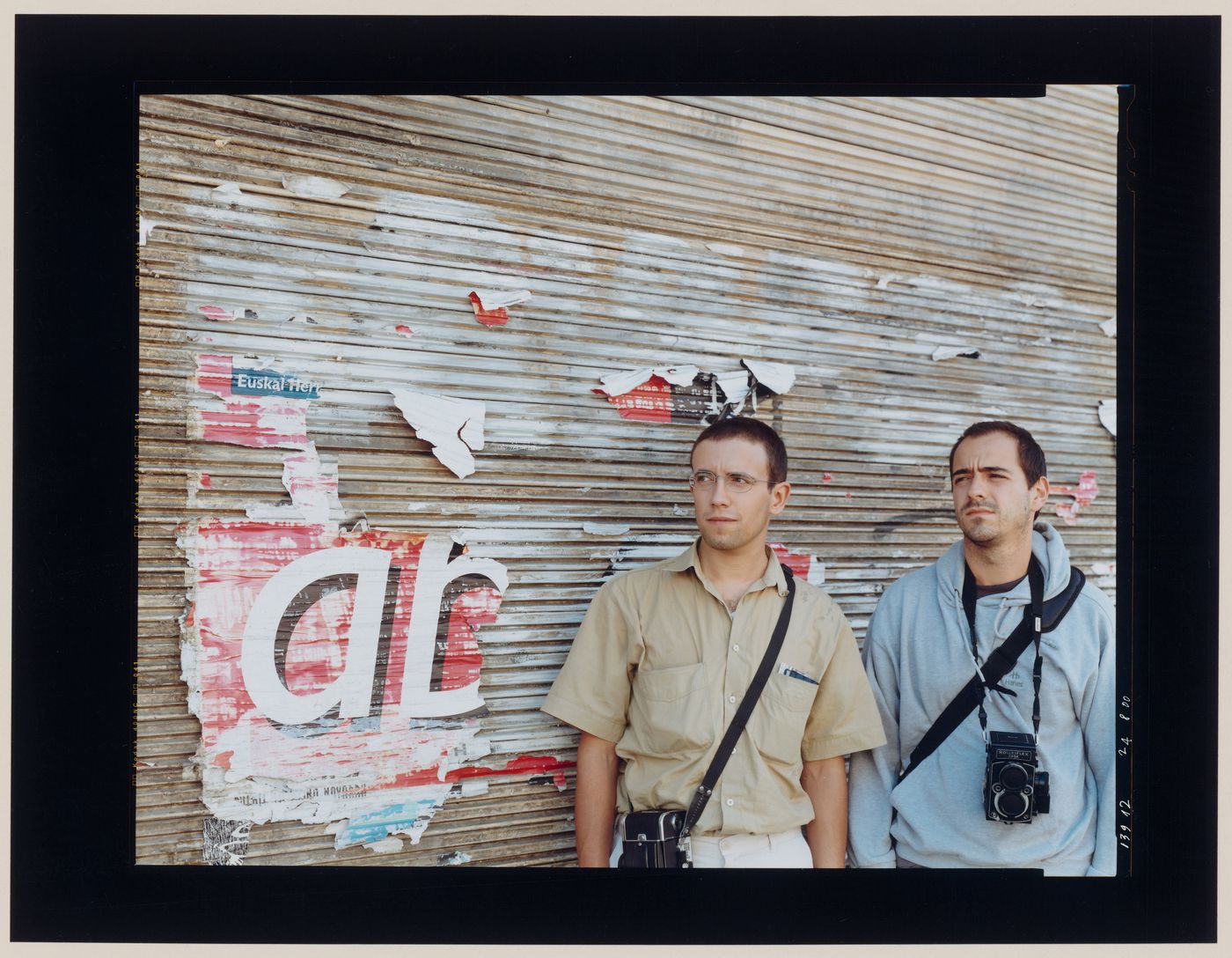 Group portrait of two men with cameras standing in front of a wall bearing graffiti and torn posters, Puente la Reina, Spain (from the series "In between cities")