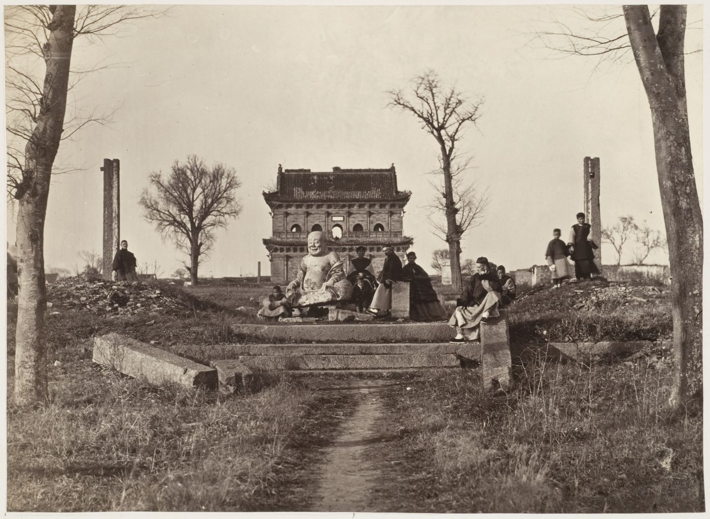 View of a group of people and a statue of Maitreya Buddha [Mi-le Fo] with the ruins of the Building for Buddhist Scripture in the background, K'ai-yuan [Kaiyuan] Temple, near Soochow (now Suzhou), China