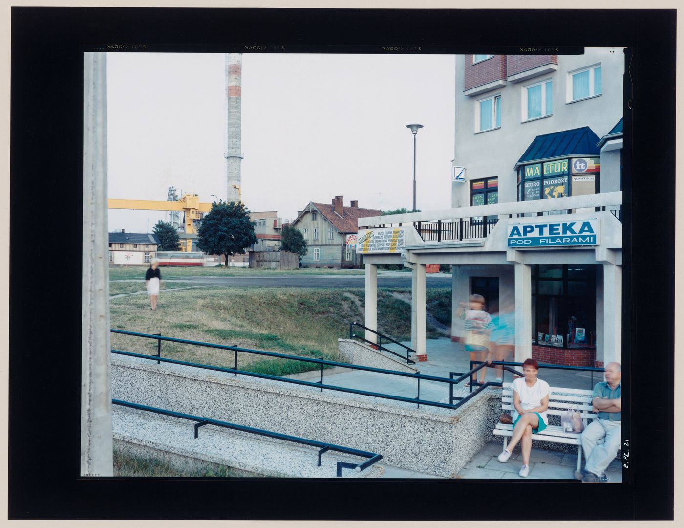 View of the entrance to a commercial building showing people in the foreground and a smokestack in the background, Malbork, Poland (from the series "In between cities")