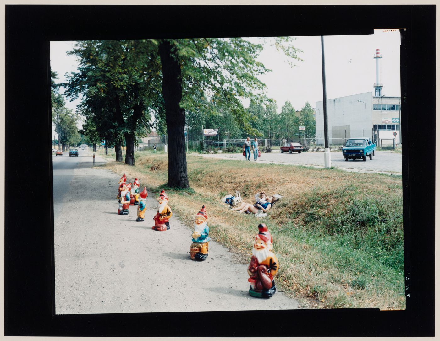 View of garden gnomes and people beside a road showing a building in the background, Cierznie, near Jastrowie, Poland (from the series "In between cities")