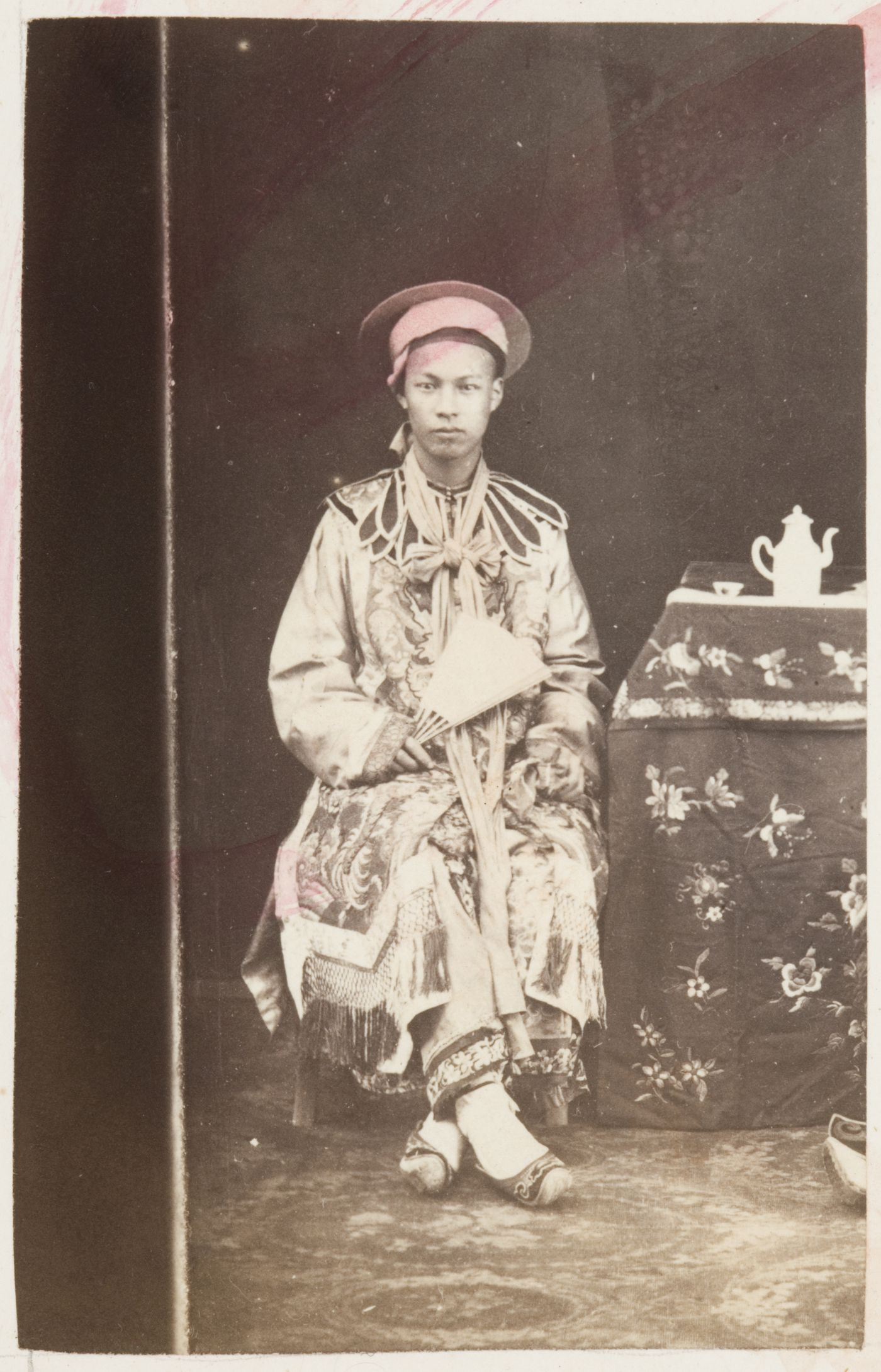 Portrait of a Chinese man seated beside a stand and holding a fan