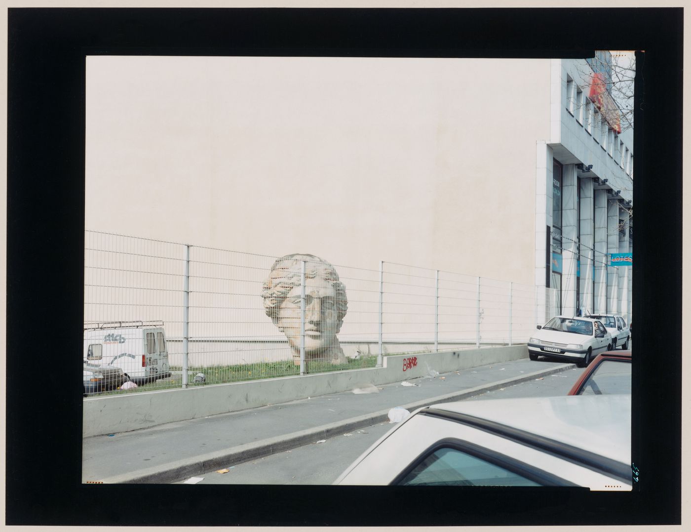 View of a sculpted head derived from the Venus de Milo showing a fence and a street in the foreground and a building in the background, Saint-Denis, France (from the series "In between cities")