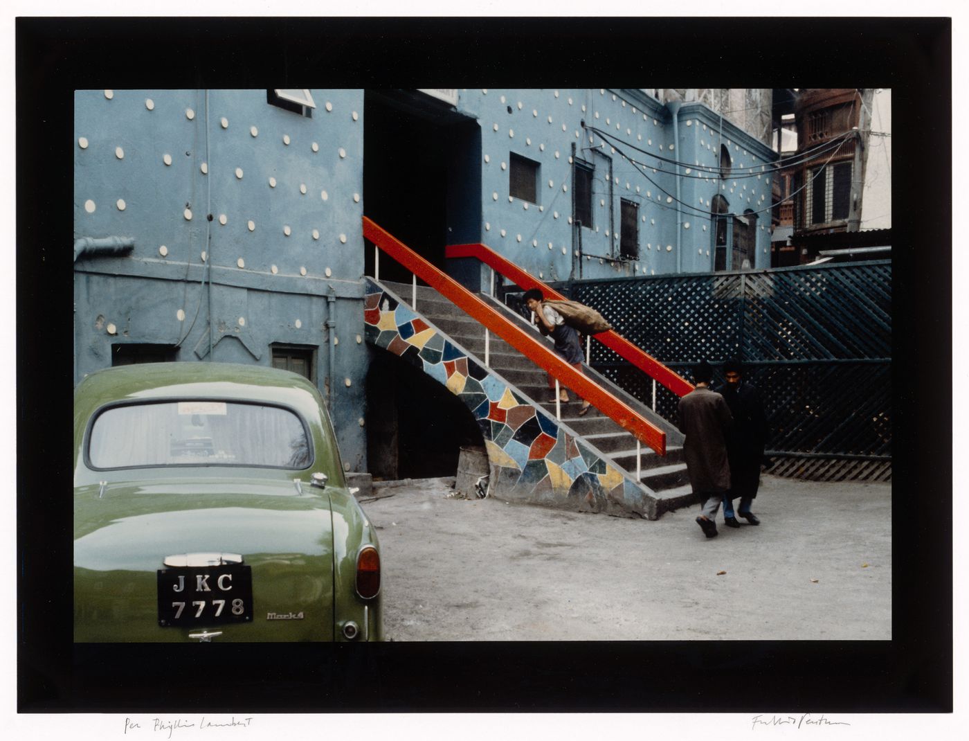 Street level view of a house painted in blue with a mosaic staircase, showing people in the background and a green car in the foreground, Srinagar, Kashmir, India