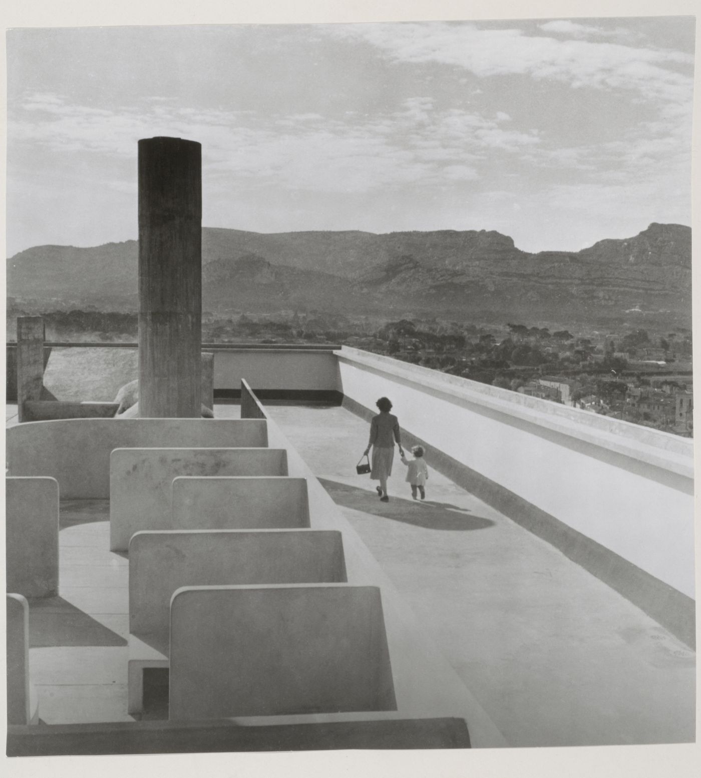 View of the roof of Unité d'habitation showing a woman and child on the running track and mountains in the distance, Marseille, France