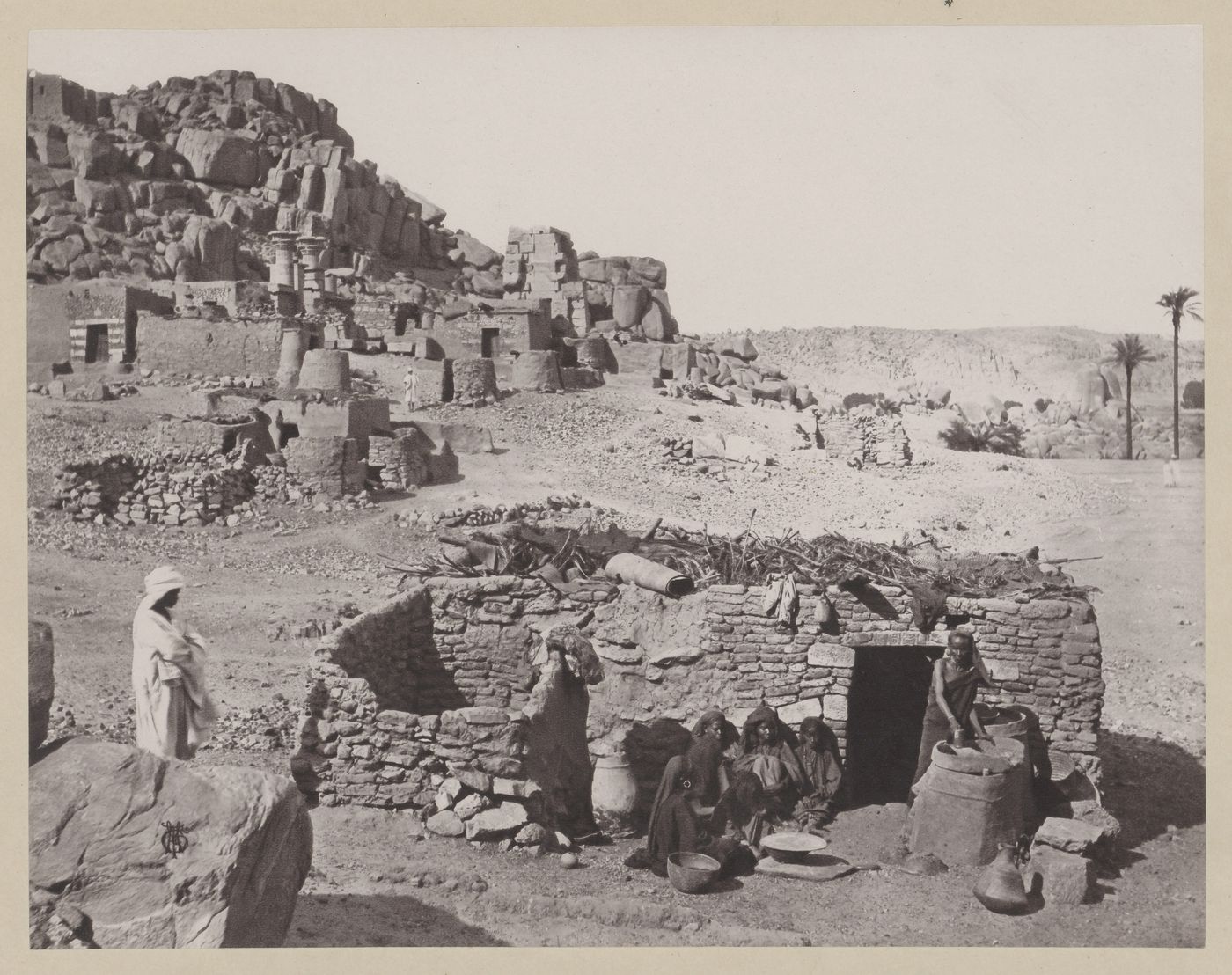 View of Nubian people in front of low stone building in the foreground and archaeological ruins in the background, Bigeh, Egypt