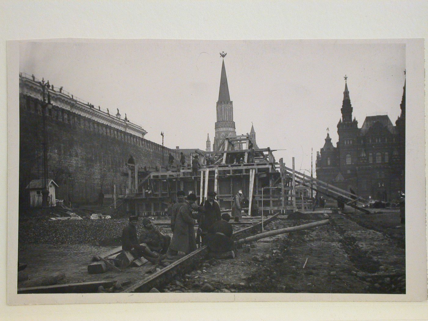 View of the lateral façade of the second wooden Lenin Mausoleum under construction showing workers installing tramway rails with Nikol'skaia Tower (Nicholas Tower) in the background and the Historical Museum on the far right, Red Square, Moscow