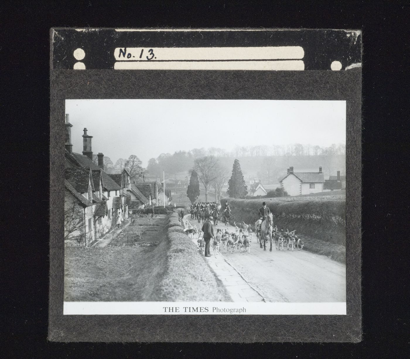 View of a fox hunt with a pack of hounds, master, huntsmen and followers riding on a road through a village in the countryside.